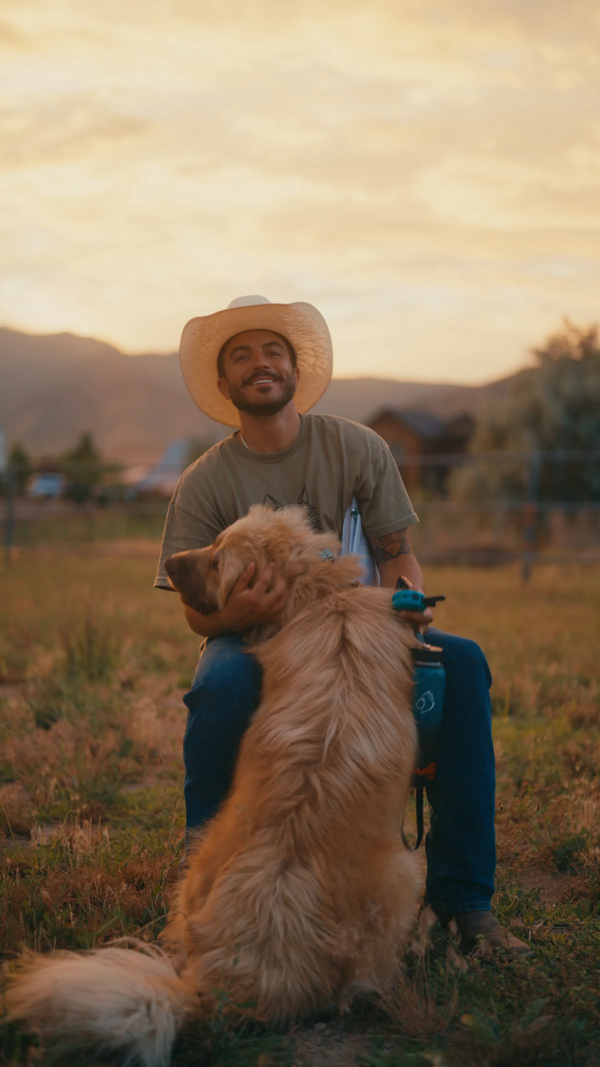 A rancher in a cowboy hat smiling with his golden retriever at sunset.
